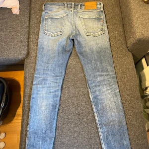  Replay Ambass Slim fit  - Snygga blå jeans från Replay med slitna detaljer och en klassisk femficksdesign. De har en knappgylf och är tillverkade i ett bekvämt denimtyg. Perfekta för en avslappnad stil.