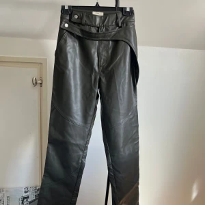 Khaki färgade skinnbyxor med knappar från A-dsgn  - Snygga svarta skinnbyxor med hög midja och stilrena knappar. Byxorna har en unik design med detaljerade sömmar och en modern passform. Perfekta för att ge din outfit en edgy touch.