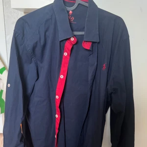 Mörkblå skjorta från Ralph Lauren - Snygg mörkblå skjorta från Ralph Lauren med kontrasterande röda detaljer och knappar. Den har en klassisk krage och det ikoniska broderade logotypen på bröstet. Perfekt för en stilren look.