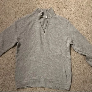 Grå tröja med dragkedja från Selected Homme - Säljer en stilren grå tröja från Selected Homme. Tröjan har en halv dragkedja framtill och är ribbad vid kragen och ärmsluten. Perfekt för en avslappnad men snygg look. Tröjan är nästan som ny!