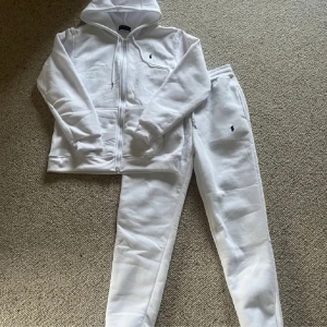 Vit hoodie och byxor från Ralph Lauren - Säljer en stilren vit hoodie med dragkedja och matchande byxor från Ralph Lauren. Hoodien har en klassisk design med fickor och en liten logotyp på bröstet. Byxorna har en bekväm passform med resår i midjan och vid anklarna. Perfekt för en avslappnad look.