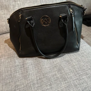 Svart handväska från Michael Kors - Snygg svart handväska från Michael Kors i skinn med guldfärgade detaljer och dragkedjor. Väskan har två handtag och en rymlig insida med en innerficka. Även en ficka på framsidan av väskan. Perfekt för att bära med sig alla nödvändigheter med stil.