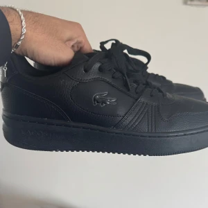 Svarta sneakers från Lacoste - Snygga svarta sneakers från Lacoste i skinn med klassisk snörning. Perfekta för en stilren look med den ikoniska krokodilloggan på sidan. Skorna har en rund tå och en bekväm sula för daglig användning. Skorna har bara används en gång, priset kan diskuteras. Köparen står för frakt!