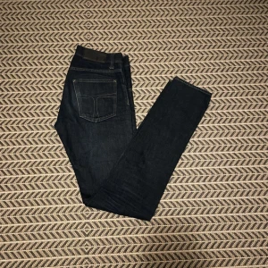 Mörkblå jeans från Tiger of Sweden - Snygga mörkblå jeans från Tiger of Sweden med klassisk femficksdesign. De har en rak passform och är tillverkade i slitstarkt denim. Perfekta för en stilren look.