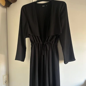Svart strand klänning från Bik Bok - Snygg svart cover on klänning från Bik Bok med långärmad design och knytning i midjan. Storlek M men passar även mindre storlekar för en lösare passform. Den aldrig kommit till användning. Perfekt för en stilren look. 