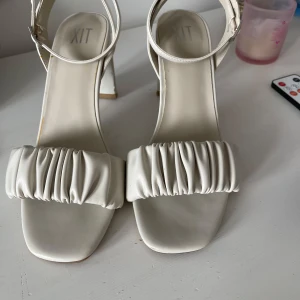 Vita klackskor från XIT - Snygga vita klackskor från XIT med öppen tå och bred klack. Skorna har en rynkad rem över foten och en justerbar rem runt ankeln. Perfekta för en stilren look. Klacken är 9 cm hög.