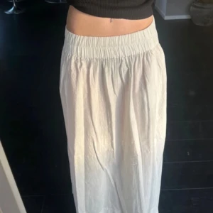 Vit långkjol med resårmidja - Säljer en stilren vit långkjol med slitt. Resår i midjan för en bekväm passform. Kjolen är perfekt för varma dagar och ger en luftig känsla. Passar bra till både enkla toppar och mer dressade överdelar.