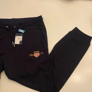 Mörkblå mjukisbyxor från Gant - Säljer ett par mörkblå mjukisbyxor från Gant med broderad logga på sidan. Byxorna har snörning i midjan och ribbade muddar vid bensluten. Små i storlek så mer som en M/L. Helt ny!