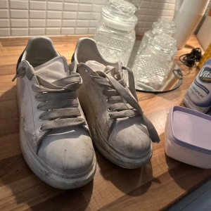  Alexander McQueen - Säljer ett par vita sneakers från Alexander McQueen med svart häl och snörning. Skorna har en chunky sula och är perfekta för en stilren look. Passar bra till både jeans och klänning.