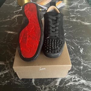 Svarta loubitins med spikar - Snygga svarta sneakers med nitar på tån och röd sula. Skorna har snörning och är tillverkade i mocka. Perfekta för att ge din outfit en edgy touch. En spik gick av