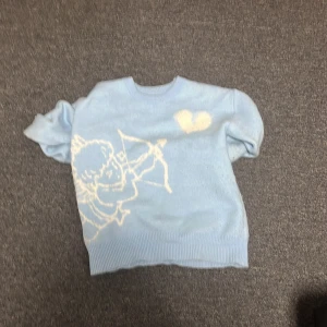 Ljusblå stickad tröja med ängelmotiv - Säljer en ljusblå stickad tröja med ett unikt ängelmotiv och texten 'I WOULD RATHER NOT LOVE' på baksidan. Tröjan har långa ärmar och ribbade kanter. Perfekt för en avslappnad stil.