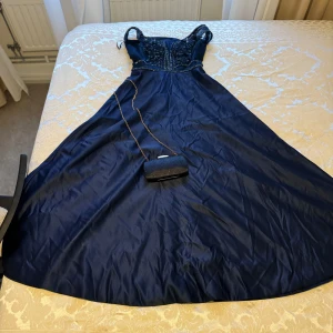 Marinblå långklänning med broderad topp - Elegant marinblå långklänning med en vacker broderad topp och pärldetaljer. Klänningen har smala axelband och en flytande kjol som ger en fin silhuett. Klänningen har en korsett som knyts bakom, vilket ger möjligheten till att välja rätt passform. Storleken går att justera från xs till M Perfekt för speciella tillfällen.