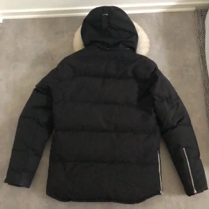 Svart dunjacka med pälsdetalj från Moose knuckles  - Säljer en svart dunjacka från Moose knuckles med avtagbar huva och pälsdetalj. Jackan har flera fickor med dragkedja och knappar framtill. Perfekt för kalla dagar!