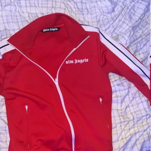 Röd kofta från Palm Angels - Säljer en snygg röd jacka från Palm Angels med vit dragkedja och vita ränder på ärmarna. Jackan har en hög krage och två fickor med dragkedja framtill. Perfekt för en sportig look! Storleken är S. Den är knappt använd för den passar inte mig! Den är äkta, men har ej kvitto för köptes av min vän!!!