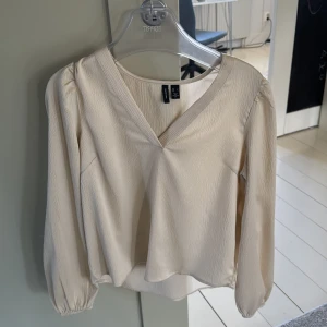 Beige blus från Vero Moda - Säljer en stilren beige blus från Vero Moda med v-ringning och långa ärmar. Blusen har en lätt textur och elastiska ärmslut för en bekväm passform. Perfekt för en elegant look.