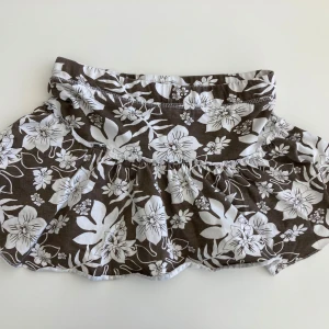 Brun blommig kortkjol - Söt brun kortkjol med vitt blommönster. Kjolen har en volangdetalj som ger den en lekfull touch. Perfekt för varma dagar.