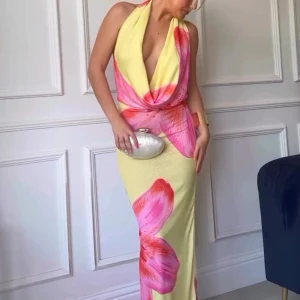 Gul och rosa långklänning med blomtryck  - Snygg långklänning i ljusgul med stora rosa blommönster. Klänningen har en djup v-ringning och är ärmlös med halterneck-design och djup rygg. Helt oanvänd med etikett kvar.