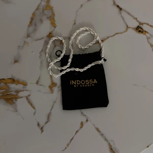Silverfärgat halsband från Indossa - Elegant silverfärgat halsband från Indossa med en vriden design. Perfekt för att ge en stilren touch till din outfit. Kommer i en snygg svart påse med guldfärgad logotyp.