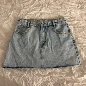 Ljusblå jeanskjol från Pull&Bear - Snygg ljusblå jeanskjol från Pull&Bear med klassisk femficksdesign och knappstängning fram. Perfekt för en avslappnad stil. Köpt för ett år sedan men har aldrig använt den. 