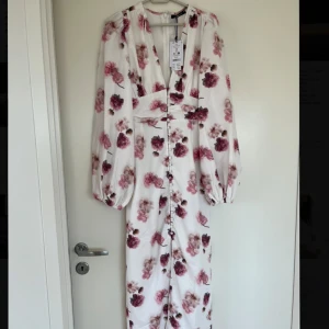 Balloon Sleeve dress Gina Tricot - Elegant vit långklänning med rosa blommönster och v-ringning. Klänningen har långa ärmar och knappar framtill samt en slits för extra stil. Perfekt för sommarens festligheter. Från Gina Tricot med lapparna kvar. 