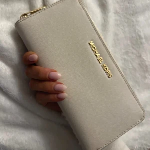 Beige plånbok från Michael Kors - Snygg beige plånbok från Michael Kors med guldfärgad dragkedja och logotyp. Plånboken har flera fack för kort och sedlar, perfekt för att hålla ordning på dina tillhörigheter. Stilren och praktisk design som passar till alla tillfällen.