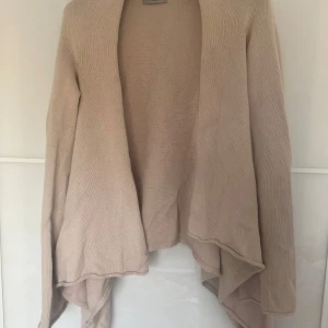 Beige stickad kofta från Hunkydory - Säljer en stilren beige stickad kofta från Hunkydory Essentials. Koftan har en öppen design med långa ärmar och ett mjukt fall. Perfekt för lager-på-lager under kyligare dagar. Passar till både vardag och mer uppklädda tillfällen.