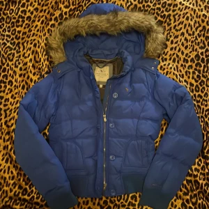 Blå dunjacka från Abercrombie - Säljer en snygg blå dunjacka/ pufferjacka från Abercrombie med pälskantad huva. Jackan har både dragkedja och knappar, men dragkedjan går inte hela vägen upp. Perfekt för kalla kvällar eller sommar jacka! Konatakta för pris 