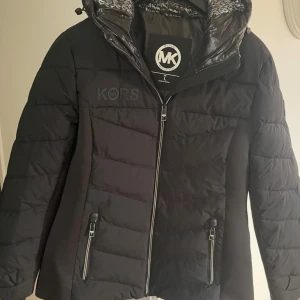 Svart pufferjacka från Michael Kors - Snygg svart pufferjacka från Michael Kors med huva och dragkedja framtill. Jackan har quiltad design, två fickor med dragkedja och diskret MK-logga på bröstet. Perfekt för kalla dagar och riktigt stilren look.