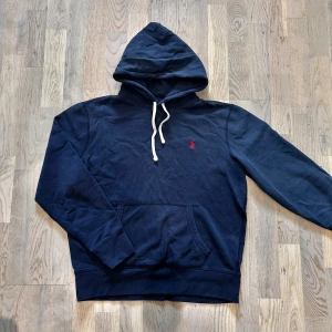 Mörkblå Ralph Lauren hoodie - Säljer en mörkblå hoodie från Polo Ralph Lauren med klassisk röd broderad logga på bröstet. Tröjan har huva med vita snören och en stor magficka framtill. Perfekt för en avslappnad stil.
