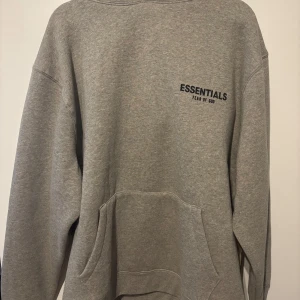 Grå Essentials Fear of God hoodie - Snygg grå hoodie från Essentials Fear of God med svart logga på bröstet och ryggen. Oversized passform, stor huva och klassisk känguruficka framtill. Perfekt för en chill och trendig streetwear-look.   Priset kan diskuteras! 