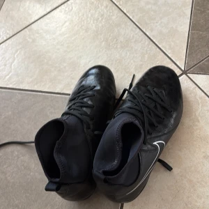 Svarta Nike fotbollsskor med dobbar - Säljer ett par svarta Nike fotbollsskor med vita detaljer och klassisk swoosh på sidan. Skorna har snörning, högre strumpliknande skaft och slitstark syntetöverdel. Yttersulan har fasta vita dobbar för bra grepp på planen. Perfekta för dig som vill ta spelet till nästa nivå. Storlek 35-36