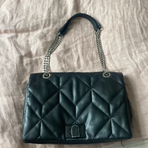 Svart  axelväska med kedja - Snygg svart axelväska i quiltad design med silverfärgad kedjerem och läderdetalj. Väskan har ett stilrent lås framtill och flera fack inuti, inklusive ett med dragkedja. 30cm bred ca 22cm höjd. Köpt på zalando men är från parfois. 