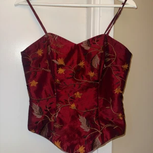 Vinröd broderad korsett med blommor - Supersnygg vinröd korsett med tunna axelband och broderade blommor i gult och brunt. Toppen har en figurnära passform, markerad midja och dragkedja bak. Materialet är glansigt och ger en lyxig känsla, perfekt för att sticka ut. Den är vintage  ( står tyvärr inget märke på den)