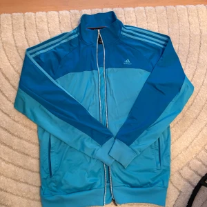 Blå vindjacka från Adidas M - Snygg blå vindjacka från Adidas med klassiska ränder på axlarna och dragkedja hela vägen. Jackan har hög krage, ribbade muddar och två fickor framtill. Perfekt för dig som gillar sportig stil och vill sticka ut med färg.