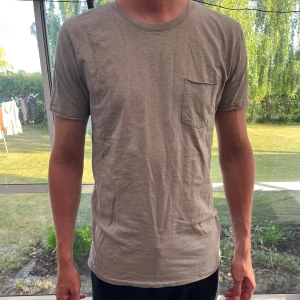 Beige basic t-shirt med bröstficka - Enkel beige t-shirt i mjuk bomull med rund halsringning och en liten bröstficka på vänster sida. T-shirten har korta ärmar och en avslappnad passform, perfekt för en chill och stilren look. Från carlings 