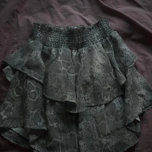 Grå volangkjol med ormmönster Gina Tricot - Supersnygg kort volangkjol från Gina Tricot i grått ormmönster. Kjolen har smock i midjan för en bekväm passform och flera lager volanger som ger en cool och trendig look. Perfekt för dig som vill sticka ut lite extra.