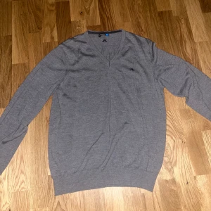 Grå v-ringad tröja J.Lindeberg - Snygg grå v-ringad tröja från J.Lindeberg med diskret logga på bröstet och nacken. Tröjan är långärmad och har en klassisk, stilren look. Perfekt att matcha med jeans eller chinos för en clean outfit. Den har krympt i tvätten så tänkte sälja den vidare