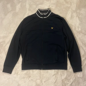 Svart track jacket från Lyle & Scott - Svart track jacket från Lyle & Scott med vit rand på kragen och klassisk gul logga på bröstet. Jackan har hel dragkedja, två fickor fram och ribbade muddar. Perfekt för en sportig och clean stil.