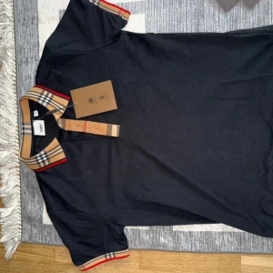 Burberry pikétröja svart med rutig krage - Snygg svart pikétröja från Burberry med klassisk beige och röd rutig krage och ärmslut. Tröjan har korta ärmar och knäppning framtill. Perfekt för dig som vill ha en stilren och lyxig look med ikoniska Burberry-detaljer.