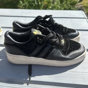 Svarta Adidas sneakers med gulddetaljer - Snygga svarta Adidas sneakers med klassiska tre ränder på sidan och guldfärgade loggor på plös och snörning. Ovandel i skinn och mocka, vit sula och svarta skosnören. Perfekta för dig som gillar streetstyle och vill sticka ut med gulddetaljer.