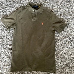 Olivgrön pikétröja från Polo Ralph Lauren - Snygg olivgrön pikétröja från Polo Ralph Lauren med klassisk krage och korta ärmar. Tröjan har orange broderad logga på bröstet och är i bomull med en skön classic fit. Perfekt för dig som gillar stilren och tidlös design. Endast använd 2 gånger. Köpt från Zalando. Nypris 1600kr. Mitt pris ENDAST 599kr!!!