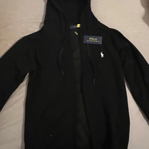 Svart hoodie från Polo Ralph Lauren - Svart kofta från Polo Ralph Lauren med klassisk broderad logga på bröstet. Tröjan har huva med snörning och dragkedja framtill. Tillverkad i mjuk bomull, perfekt för en chill och stilren look.