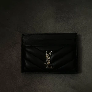 Svart YSL quiltad korthållare i skinn - Säljer en svart quiltad korthållare från YSL i äkta skinn med ikoniskt guldfärgat YSL-logo framtill. Stilren och smidig design med flera kortfack på både fram- och baksidan. Perfekt accessoar för dig som gillar lyxiga detaljer.