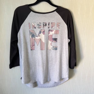 Grå och svart tröja H&M "Inspire Me" - Snygg grå tröja från H&M med svarta ärmar och texten "INSPIRE ME" framtill. Bokstäverna har ett blommigt mönster i rosa och blå toner. Tröjan har trekvartsärm och är gjord i mjuk bomull, perfekt för en chill och inspirerande look.