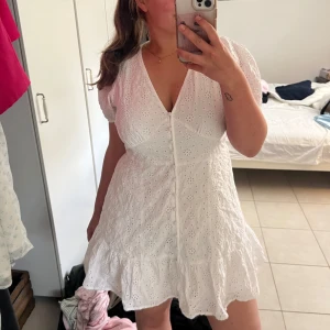 Vit kort klänning från Gina Tricot - Supersöt vit kort klänning från Gina Tricot med broderade blommor, v-ringning och knappar framtill. Puffiga korta ärmar och volangkant nertill ger en romantisk vibe. Perfekt för varma dagar och sommarhäng!