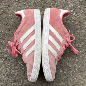 Adidas Gazelle rosa  - Säljer ett par Adidas Gazelle sneakers i rosa mocka med vita detaljer och klassiska tre ränder på sidorna. Skorna har snörning, vit sula och rosa innerfoder. Perfekta för dig som vill ha en sportig och trendig look med retrovibbar.
