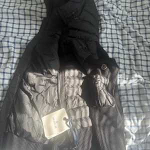 Svart Moncler pufferjacka M - Säljer en svart pufferjacka från Moncler i storlek M. Jackan har quiltade partier, stickade ärmar och detaljer, samt dragkedja framtill. Klassisk Moncler-logga på ärmen och riktigt snygg urban vibe. Perfekt för dig som vill ha både stil och värme.