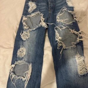 Blåa ripped jeans med stora hål - Säljer ett par blåa jeans med riktigt stora och slitna hål framtill. De har en cool, trashad look med mycket fransar och slitningar längs hela benen. Perfekta för dig som gillar streetstyle och vill sticka ut.byxorna är i bra skick 