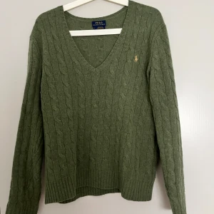Olivgrön stickad tröja Polo Ralph Lauren - Kabelstickad olivgrön tröja från Polo Ralph Lauren med v-ringning och diskret broderad logga på bröstet. Tröjan är långärmad och har ribbade muddar samt nederkant. Perfekt för dig som gillar klassisk och stilren look.