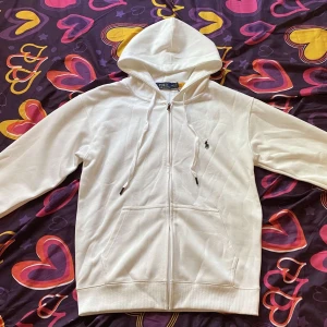 Vit hoodie från Polo Ralph Lauren M - Vit hoodie från Polo Ralph Lauren med dragkedja, snörning i huvan och klassisk broderad logga på bröstet. Tillverkad i mjuk bomull, har känguruficka och lång ärm. Perfekt för en clean och stilren look. Det är storlek S det står M men den är s i storlek. Jag säljer den för den var för liten.